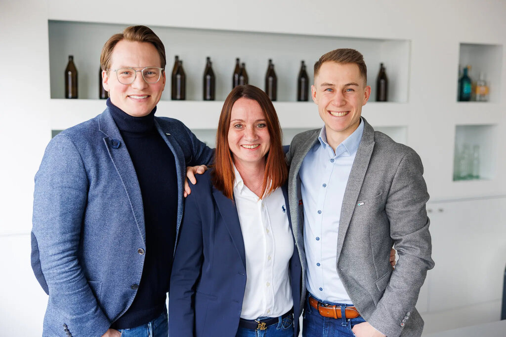 Drei Mitarbeitende aus dem Vertrieb und der Führungsebene, zwei Männer und eine Frau, posieren lächelnd in einem modernen Büro.