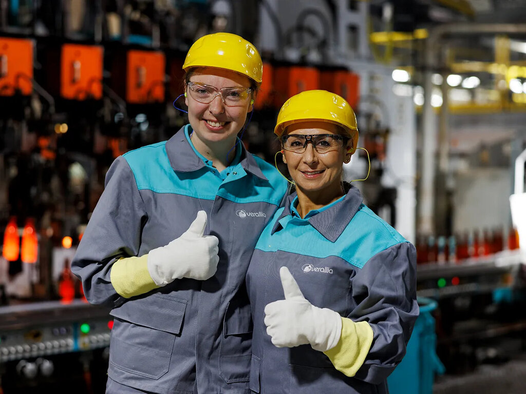Verallia-Mitarbeiterinnen in Arbeitskleidung und gelben Helmen zeigen den Daumen hoch in einer Produktionsanlage.
