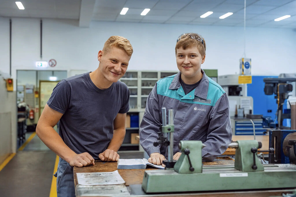 Zwei männliche Azubis in der Ausbildungs-Werkstatt, lächelnd, einer in Arbeitskleidung mit Verallia-Logo.