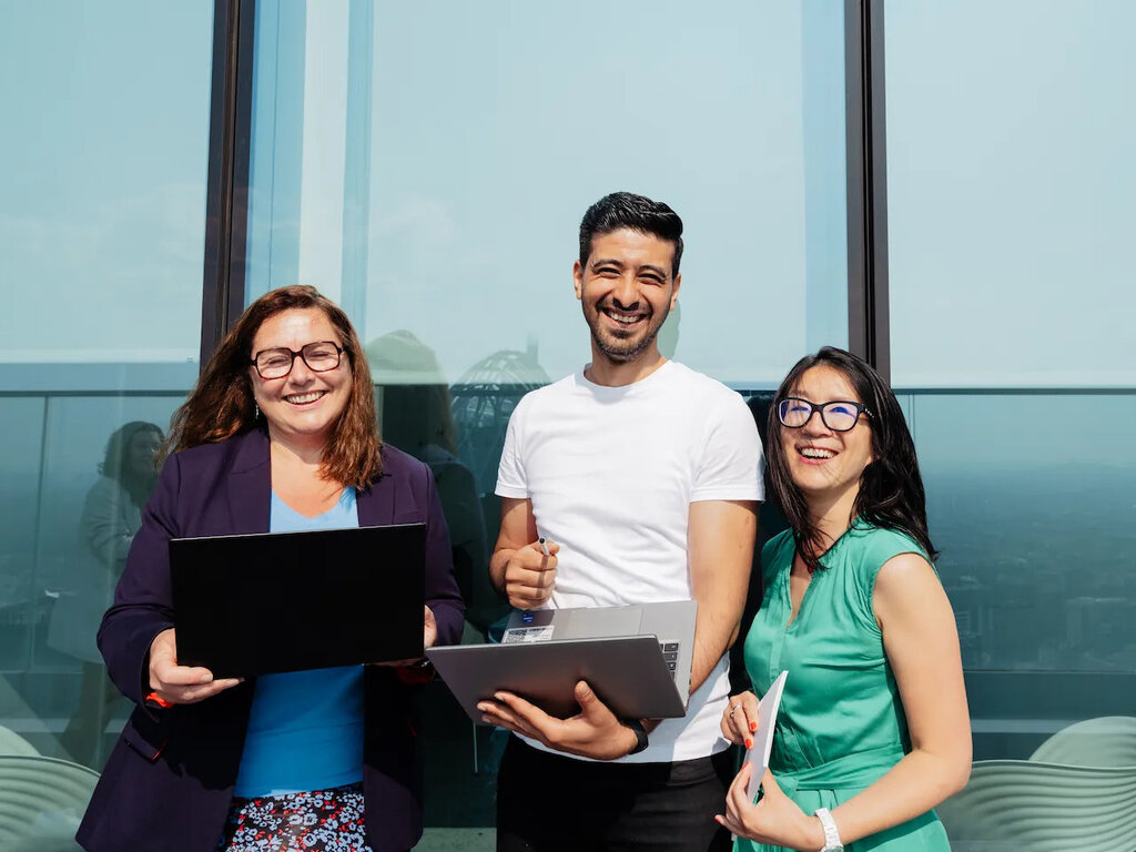 Drei Verallia-Mitarbeitende lächelnd mit Laptops und Dokumenten auf einer Terrasse mit Glasfassade.